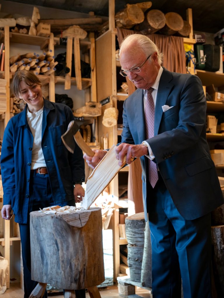 King Carl XVI Gustaf of Sweden splitting wood with an axe at Kalthoffs yxor in Stockholm while Julia Kalthoff laughs beside him