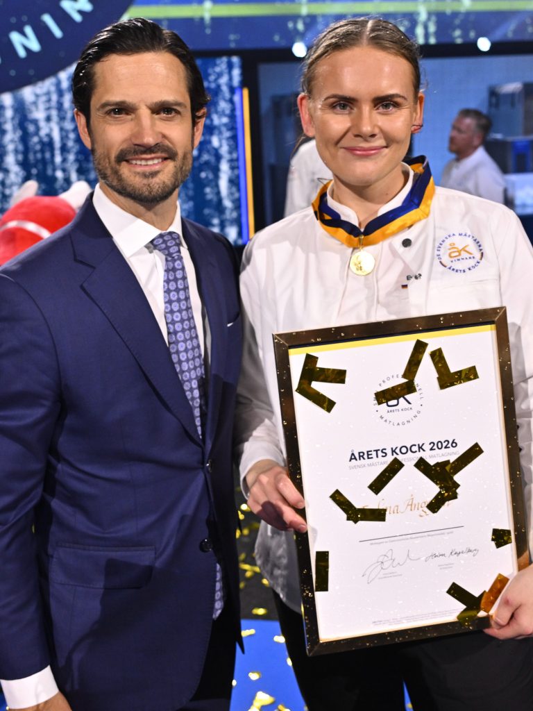 Evelina Anggren in a white chef jacket holding the framed Chef of the Year 2026 diploma beside Prince Carl Philip as gold confetti falls at Annexet in Stockholm
