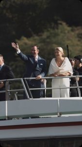 Grand Duke Guillaume waving in a navy suit with pink tie and Grand Duchess Stephanie smiling in white on the deck of the M.S. Princesse Marie-Astrid on the Moselle. Photo: Maison du Grand-Duc via @courgrandducale.