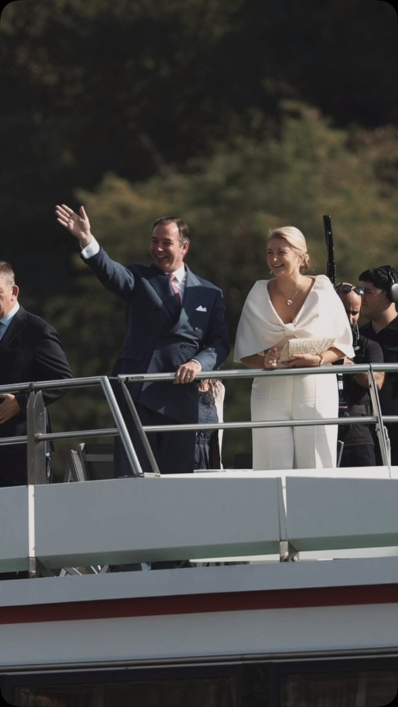 Grand Duke Guillaume waving in a navy suit with pink tie and Grand Duchess Stephanie smiling in white on the deck of the M.S. Princesse Marie-Astrid on the Moselle. Photo: Maison du Grand-Duc via @courgrandducale.