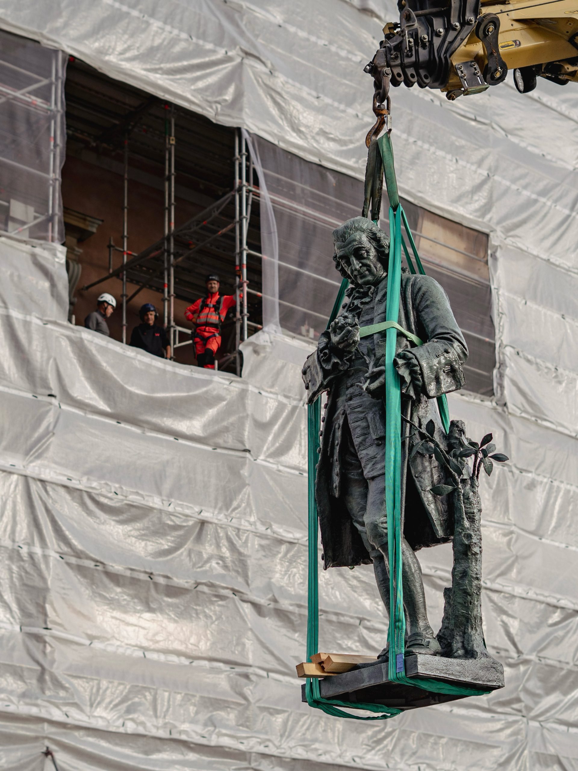 A restored dark bronze-colored statue of Carl von Linne suspended in green crane straps before Stockholm's Royal Palace facade, wrapped in white sheeting with scaffolding and workers in helmets.